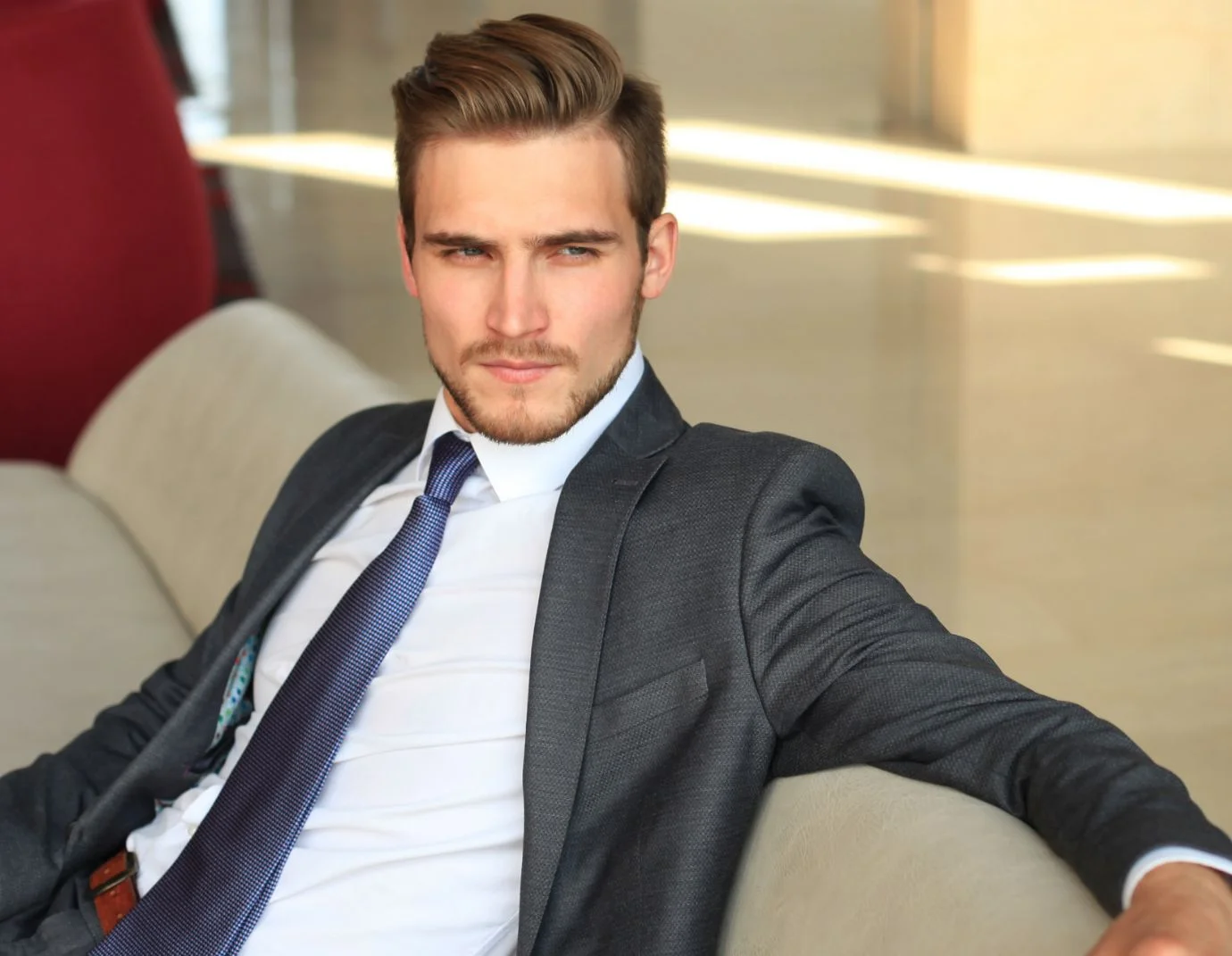 a young man sitting on a couch wearing a white shirt, grey jacket and a blue tie